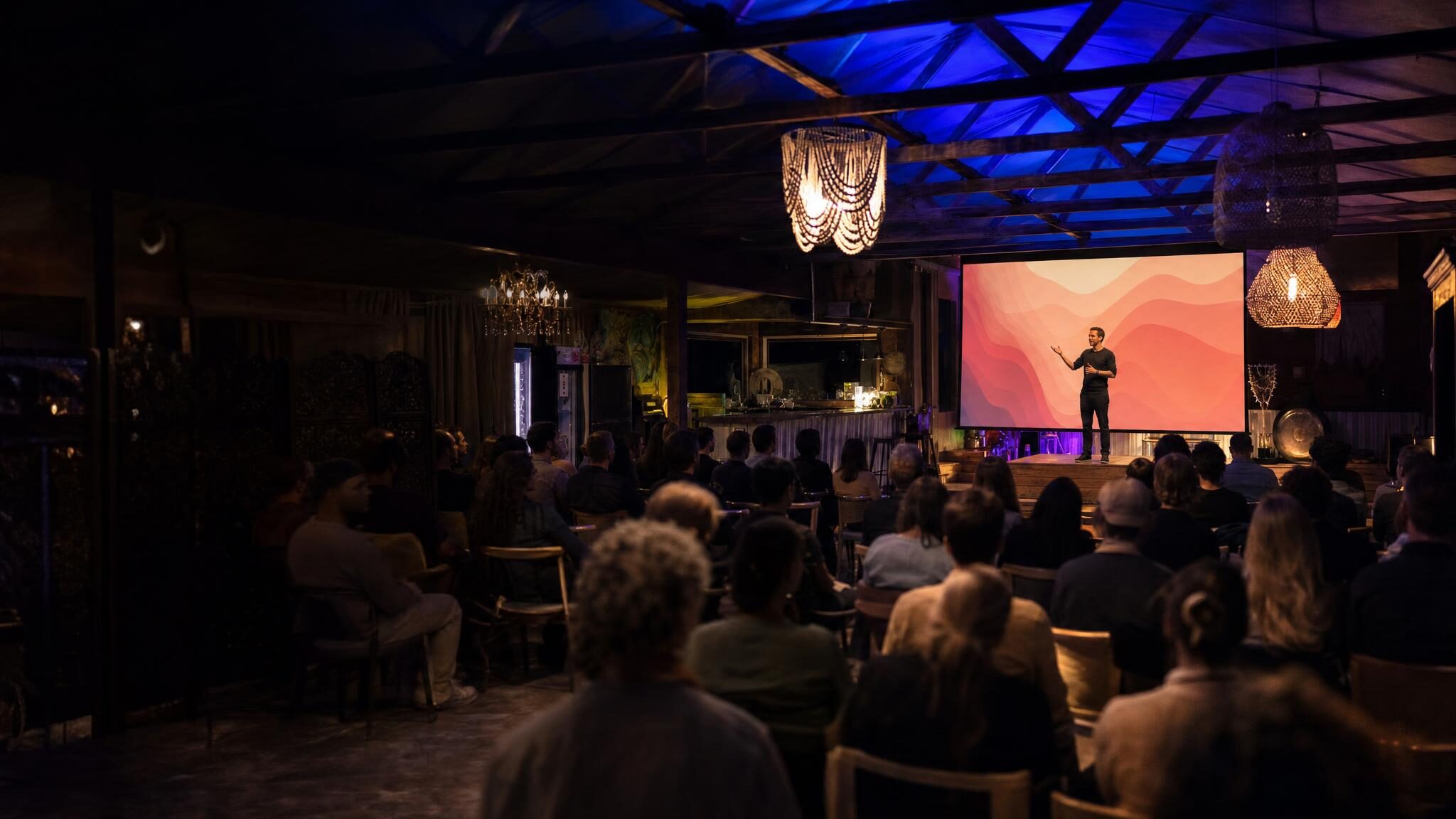 A speaker gives a keynote in front of a glowing screen while a seated audience listens inside a lounge at AWKN Ranch