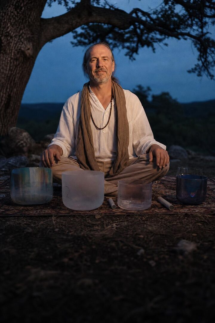 A healer seated beside singing bowls under an oak tree at dusk